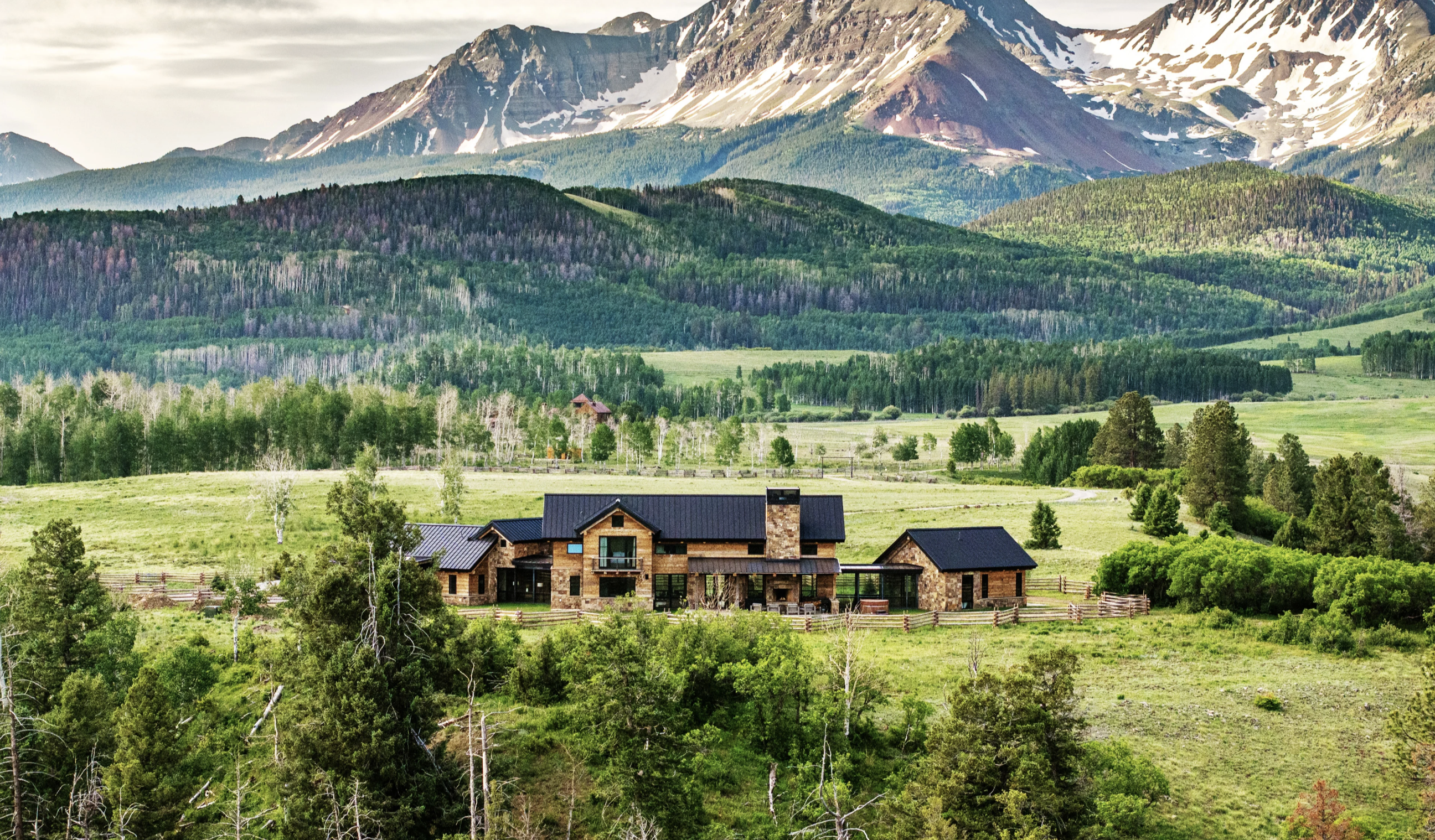Mountain home surrounded by green meadows and forest beneath snowy peaks
